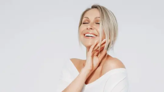 Smiling blonde woman gently touches her neck with her eyes closed