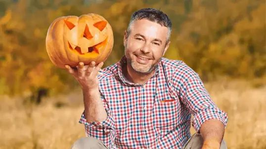A smiling man sits in a field of wheat smiling, holding a jack-o-lantern